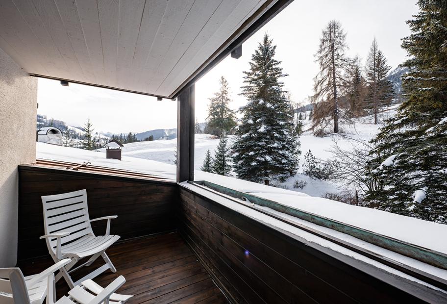 Balcone innevato con sedie, vista su alberi e montagne.